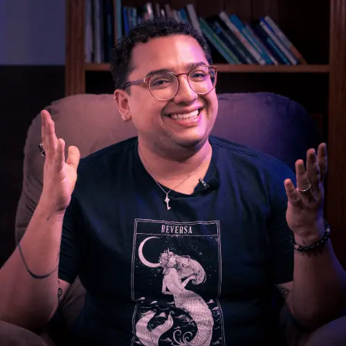 Victor Augusto sorrindo, sentado em uma poltrona diante de uma estante de livros